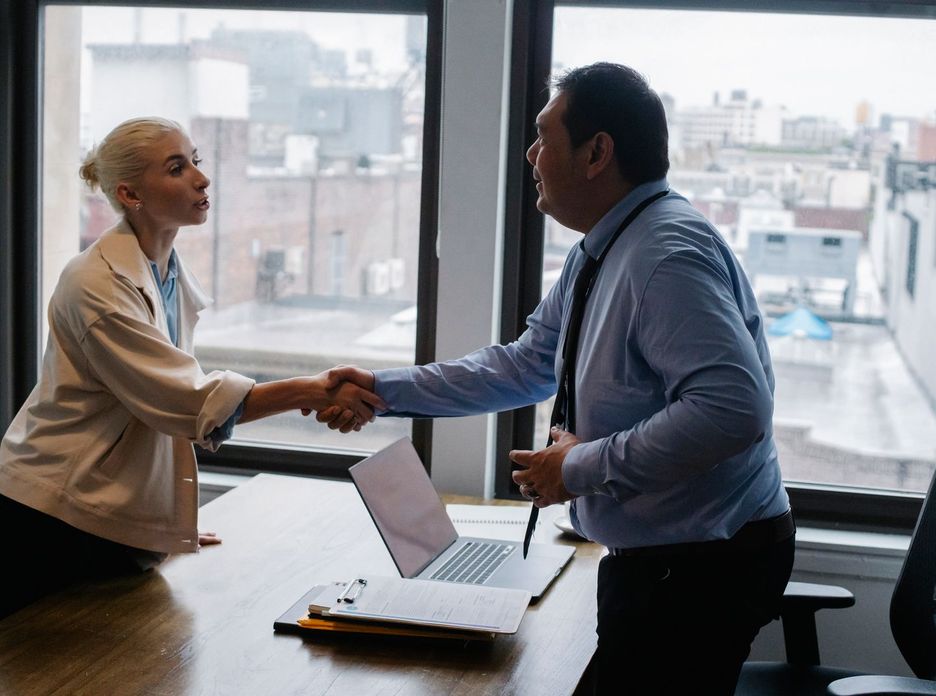 Young female candidate shaking hands with employer before job interview in light office with urban landscape through windows