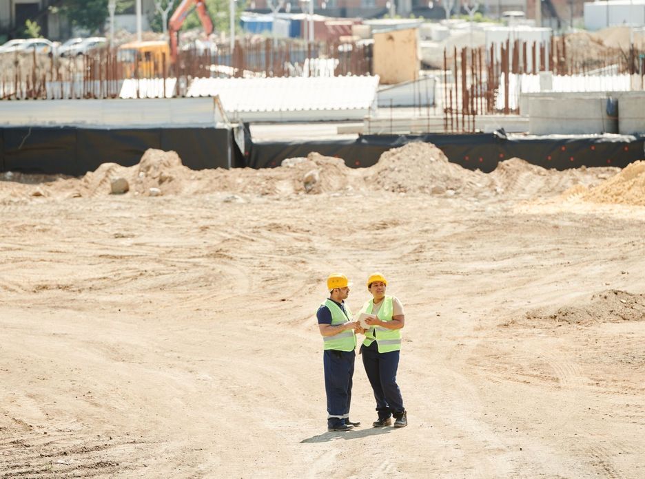 Two workers discussing plans on a sandy construction site, wearing safety gear