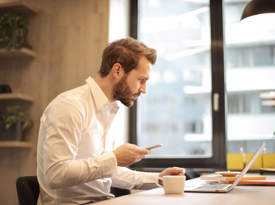 Focused businessman working on laptop while checking smartphone in modern office