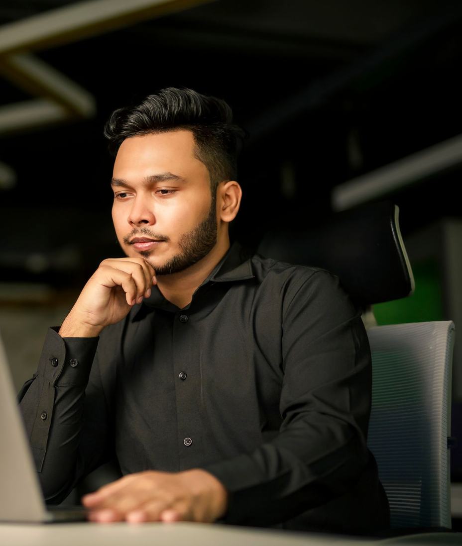 A man in a black shirt working at a laptop in a dim office, hand on his chin