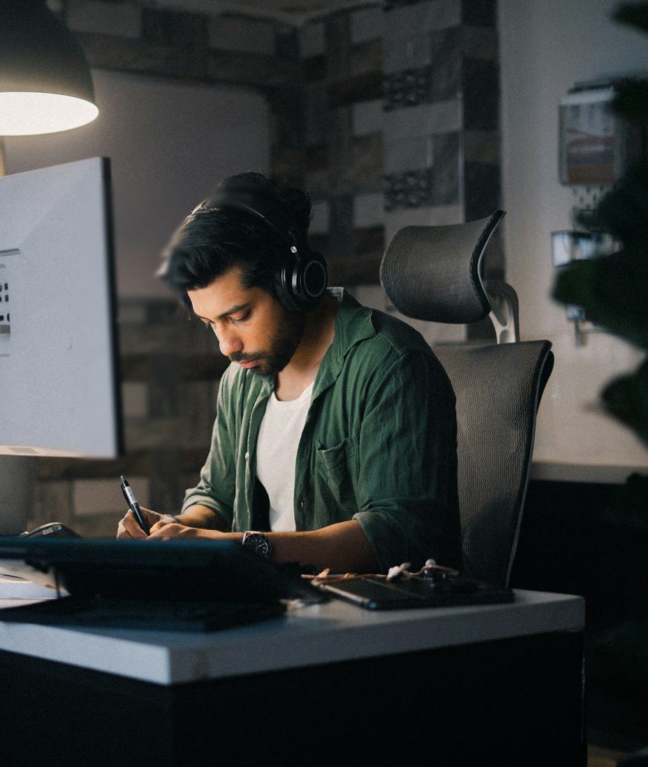 A man in headphones writing notes at his desk beside a computer monitor